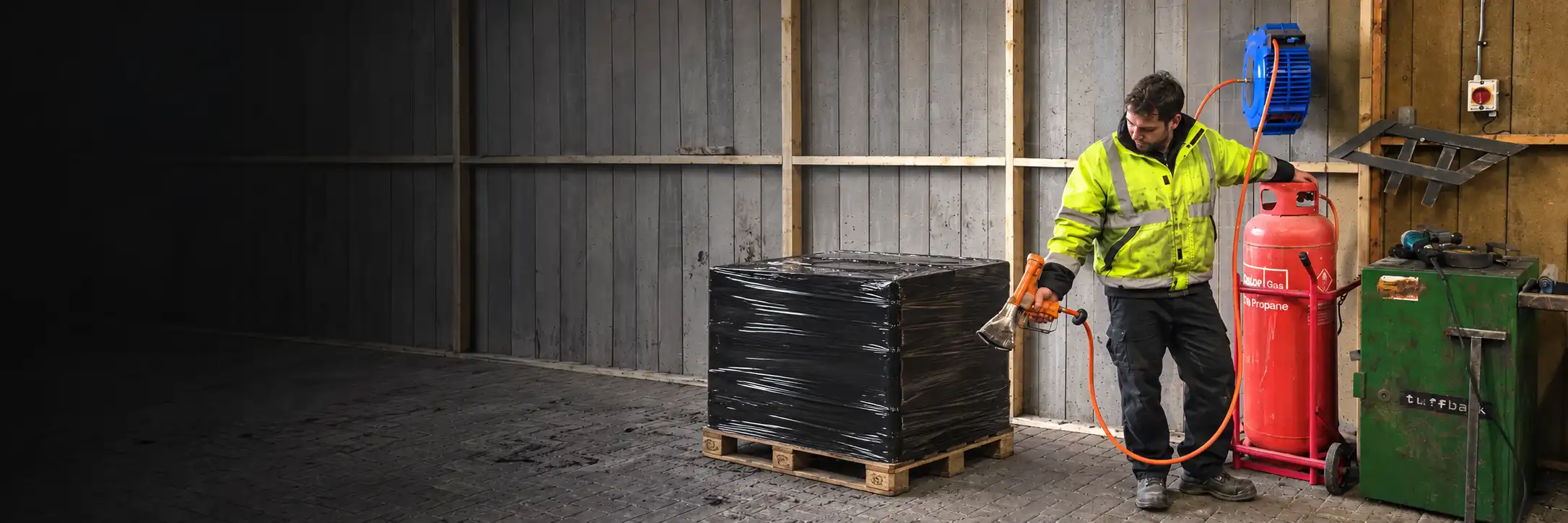 ReCoila propane hose reel and operator holding a shrink-wrap gun beside a pallet wrapped in black polythene